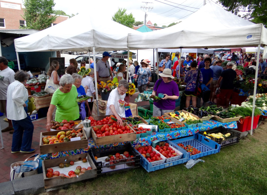 Shoppers crowd the Bath Farmers' Market under white tents, browsing tables filled with fresh tomatoes, squash, eggplant, and greens on a summer day, surrounded by local vendors and colorful produce displays.
