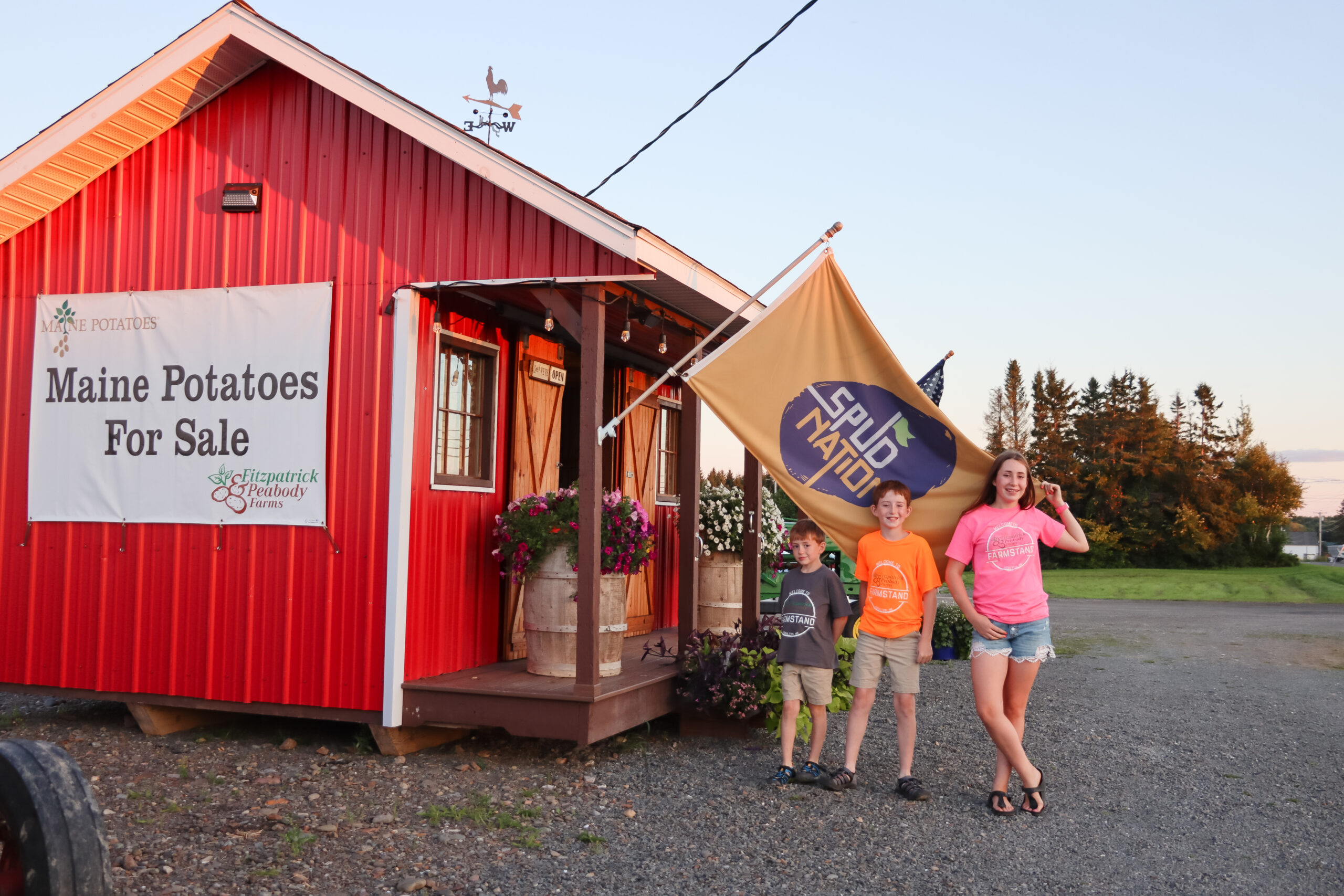 Three kids, ages 13, 11 and 8 stand in front of their farmstand where they sell potatoes, raspberries, tomatoes, and other fresh produce.