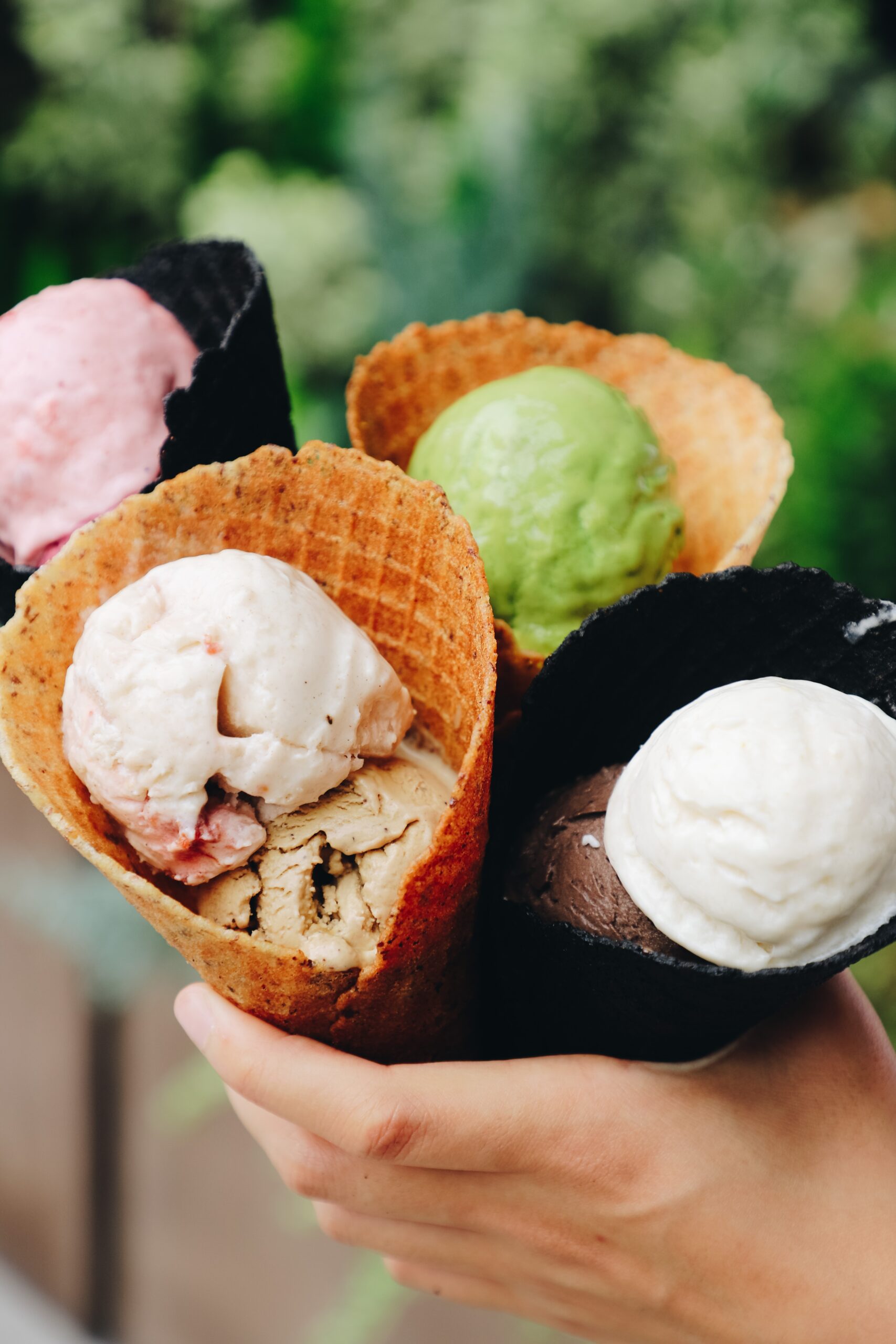 Ice Cream Cones Hand holding four waffle cones filled with vibrant, assorted ice cream scoops in a sunny outdoor setting.