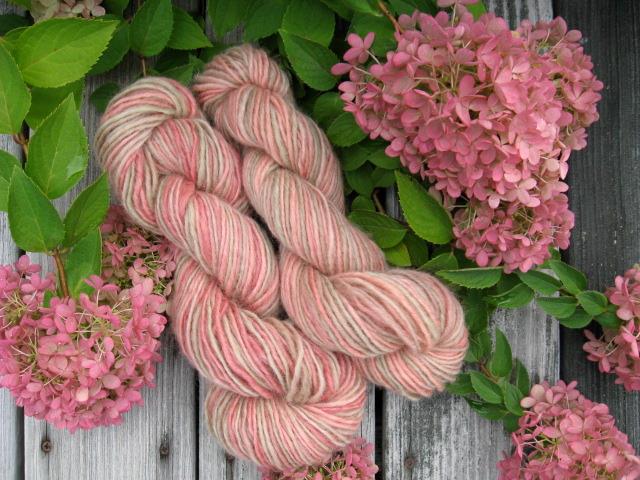 Hilltop handspun yarn hydrangea Two skeins of hand-spun pink and cream yarn displayed on weathered wood surrounded by blooming pink hydrangea flowers.
