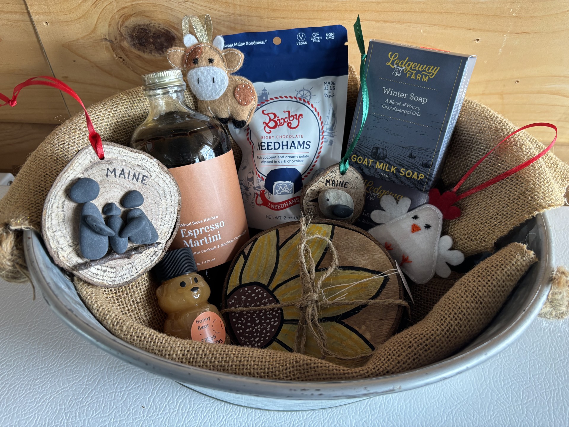 Pine Ridge Acres 4 Maine-made gift collection neatly arranged in a metal tin, including espresso martini from Wood Stove Kitchen, goat milk soap from Ledgeway Farm, Bixby Needhams chocolate, local honey, handcrafted ornaments, sunflower artwork, felt animal decorations, and ground beef coupons.